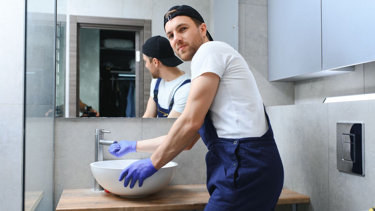 Plumber in overalls and gloves fitting a basin in a gray tiled bathroom